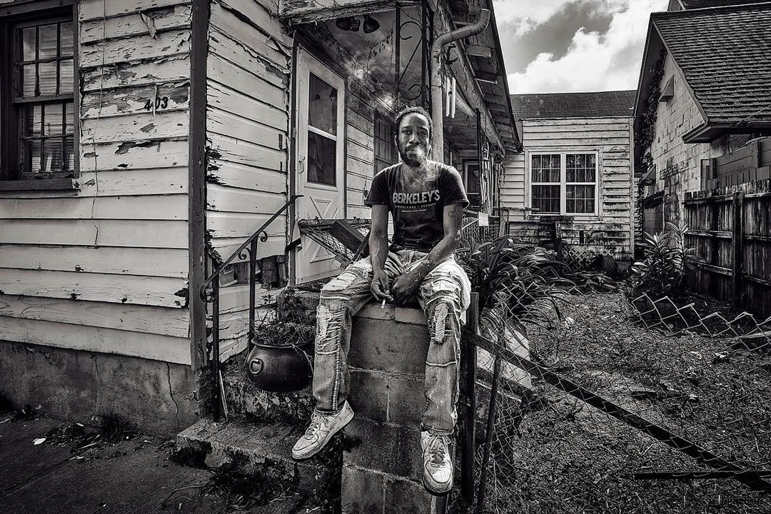 Monochrome portrait of a stylish African-American man sitting on porch of weathered house, wearing t-shirt, patched jeans, and sneakers.