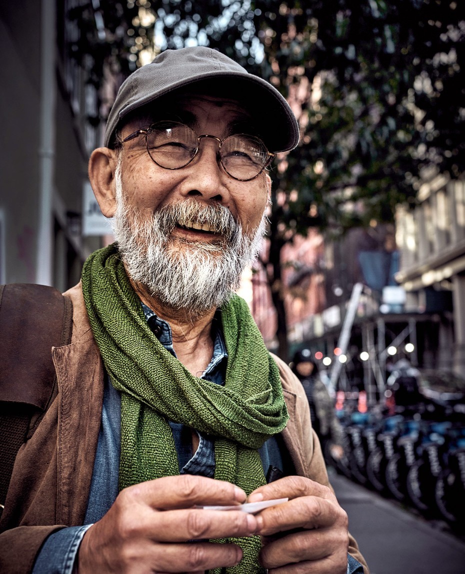 Elderly man with white beard wearing round glasses, green scarf, and grey cap outdoors on a city street
