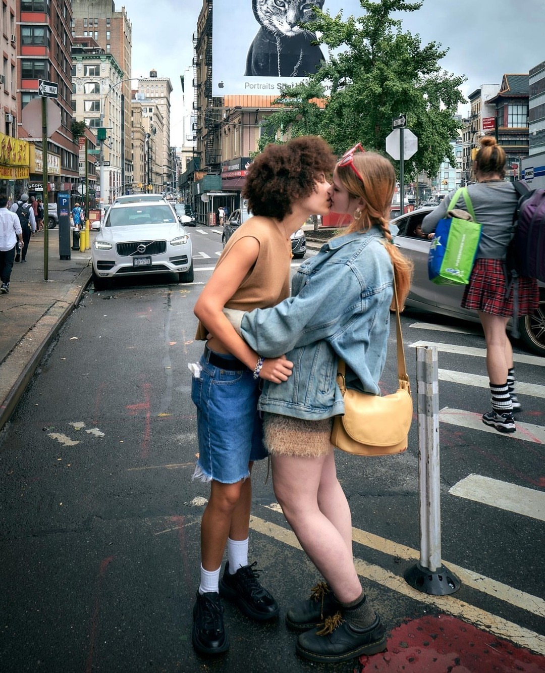 Two women embracing and kissing on a city street with buildings, traffic, and an advertisement in the background, capturing an intimate moment.