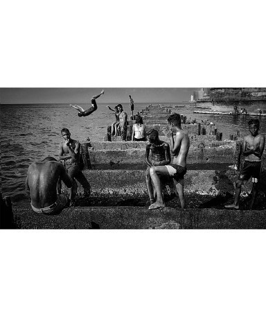Monochrome capture of a group of people near the ocean, with one diving into the water; stone steps and pilings along the coast.