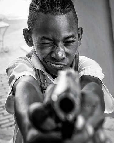 Dramatic black and white portrait of a young African male pointing a handgun directly at the viewer with a shallow depth of field.