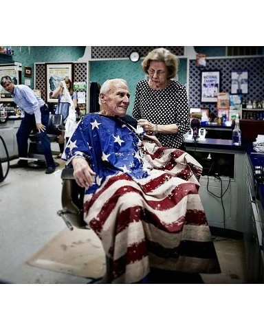 Elderly man getting a haircut in a barbershop, wearing a cape resembling the American flag, with other people in the background.
