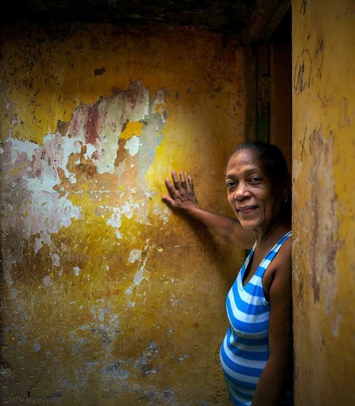 Portrait of an adult woman in a blue-and-white striped top standing in a weathered doorway, her hand resting on a yellow wall.