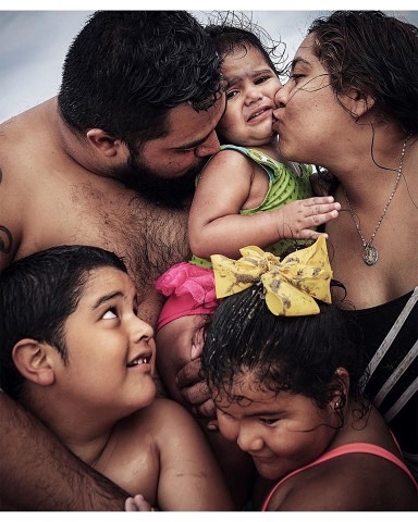 Close-up of a family group with parents kissing a crying toddler girl. Two children look on; indoor lighting.