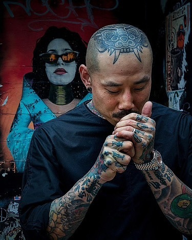 Tattooed man with shaved head lights a cigarette outdoors, mural art in background, wearing black shirt, silver ring, watch.