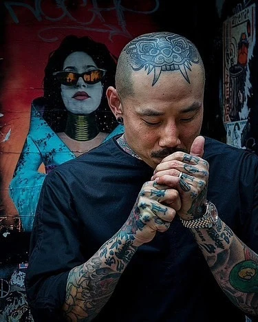 Tattooed man with shaved head lights a cigarette outdoors, mural art in background, wearing black shirt, silver ring, watch.