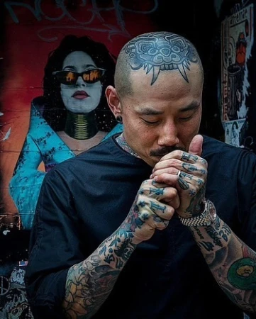Tattooed man with shaved head lights a cigarette outdoors, mural art in background, wearing black shirt, silver ring, watch.