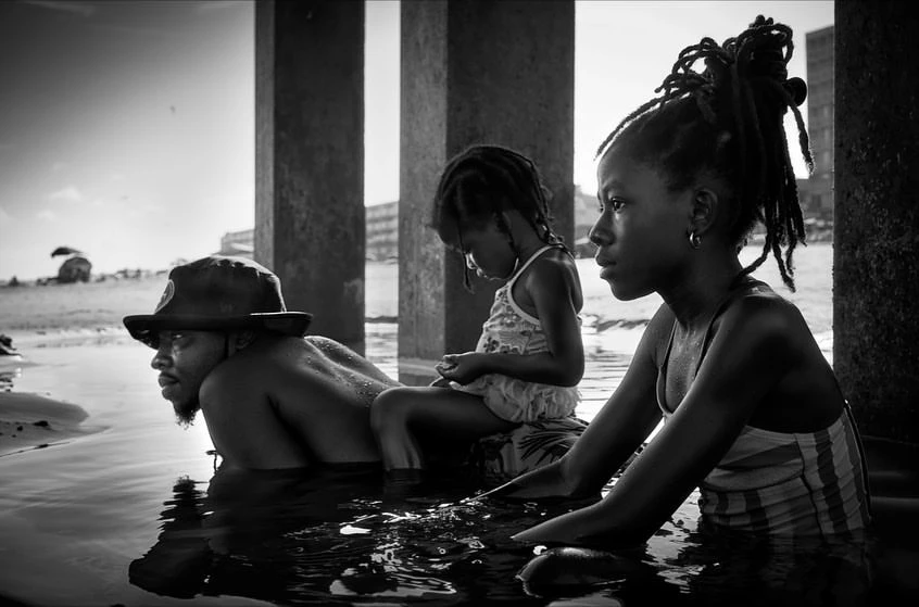 Black and white photo of an African-American family wading in shallow water. A man, girl, and woman are near concrete pillars.