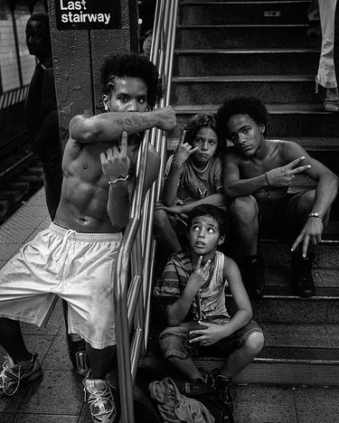 Black and white photo of shirtless young adult man standing with several children on stairs in urban environment making hand gestures.