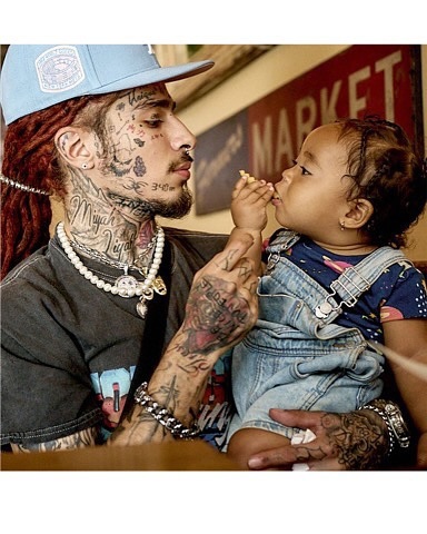 Man with tattoos sharing food with baby in overalls, indoors. Close up of the pair interacting against a market background.