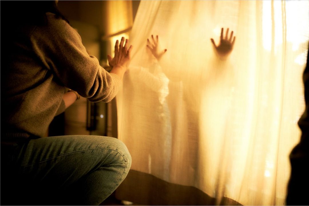 A person in a sweater and jeans interacts with two figures behind a translucent curtain in a dimly lit interior space.