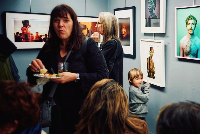 Indoor gathering with diverse people at an art gallery, viewing framed portraits of faces and figures on a grey wall, eating and drinking.