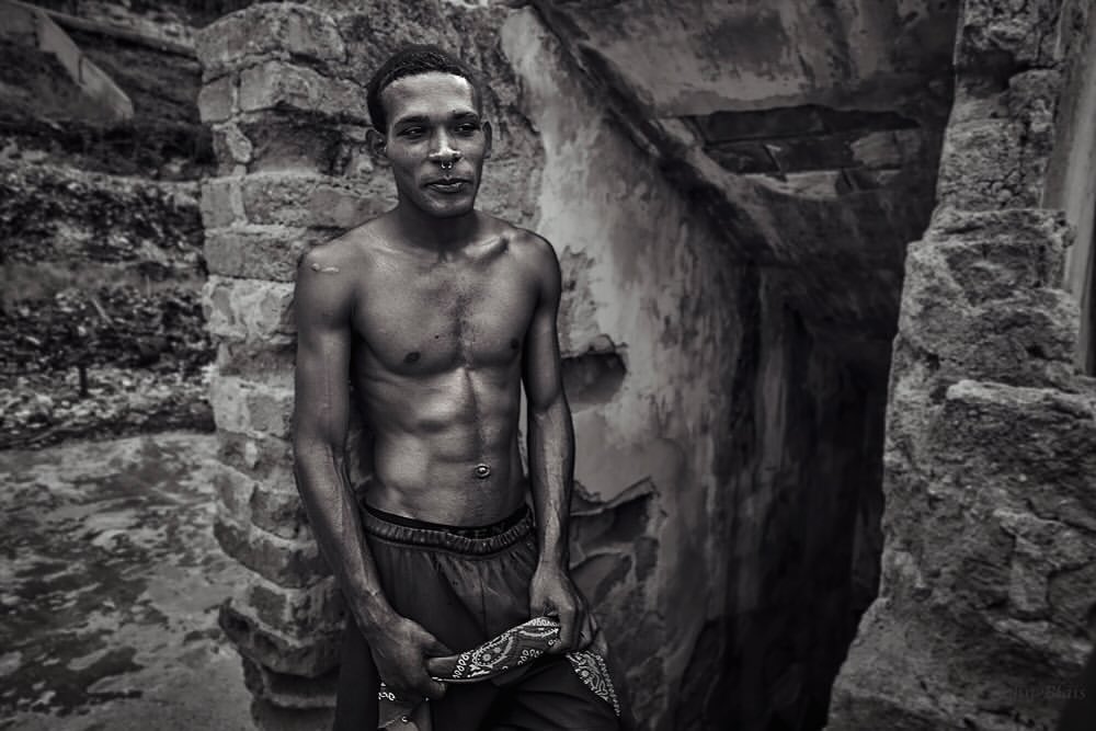 Black and white portrait of a shirtless adult male with visible abs, in an outdoor setting with crumbling stone ruins.