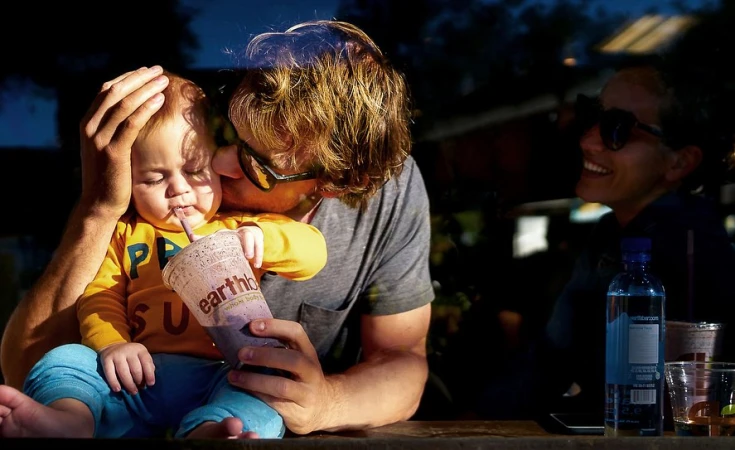 A father holds his young child who sips a smoothie, with a woman smiling in the background, creating a heartwarming family moment.