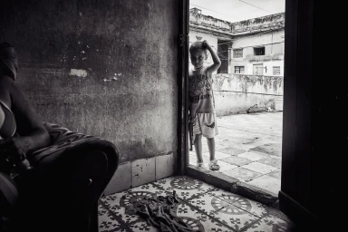 Monochrome documentary photograph by John Blais of a young child standing in an open doorway looking towards the camera.
