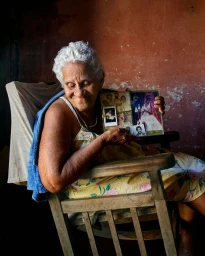 Elderly woman with white hair smiling while holding an open photo album in a wooden rocking chair inside a rustic room