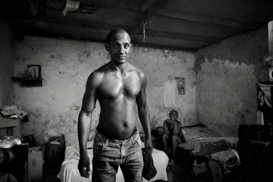 Monochrome portrait of a shirtless man standing in a rustic room with a seated woman in the background.