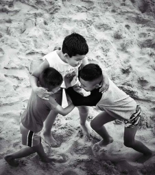 Three young boys wrestling and playing together in the sand on a beach in a high angle black and white shot