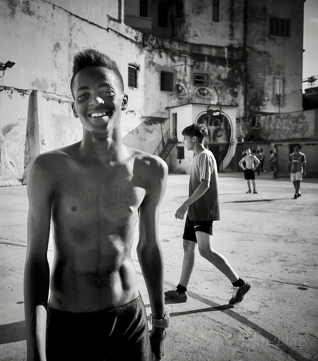 Monochrome portrait of a smiling young black man shirtless outdoors with other figures in an urban yard by John Blais