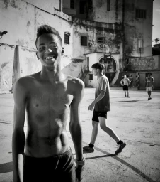 Monochrome portrait of a smiling young black man shirtless outdoors with other figures in an urban yard by John Blais