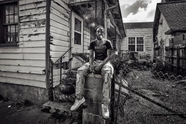 Black man sitting on concrete steps in front of a rustic wooden house, holding a cigarette and looking forward.