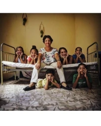 Portrait of an older woman seated center on a metal bedframe surrounded by seven family members in a domestic interior.