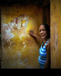 An older woman in a blue and white striped tank top touching a weathered yellow wall, photographed by John Blais.