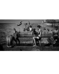 Black and white photograph by John Blais showing several young men gathered along a concrete sea wall while another dives into the water.