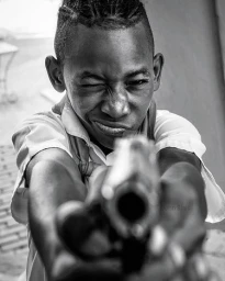 Close-up black and white portrait of a young boy making a playful face while pointing a toy gun directly at the camera.