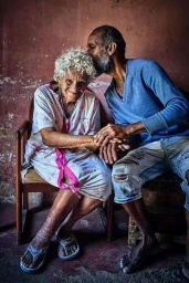 Elderly woman with white curly hair seated in a wooden chair holding hands with a bearded man kissing her head by John Blais