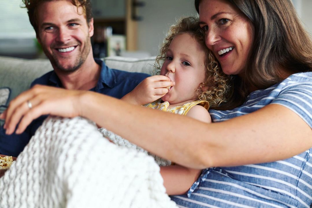 Smiling family—mother, father, and daughter—sitting together on a couch with a white blanket, creating a cozy and heartwarming indoor scene.