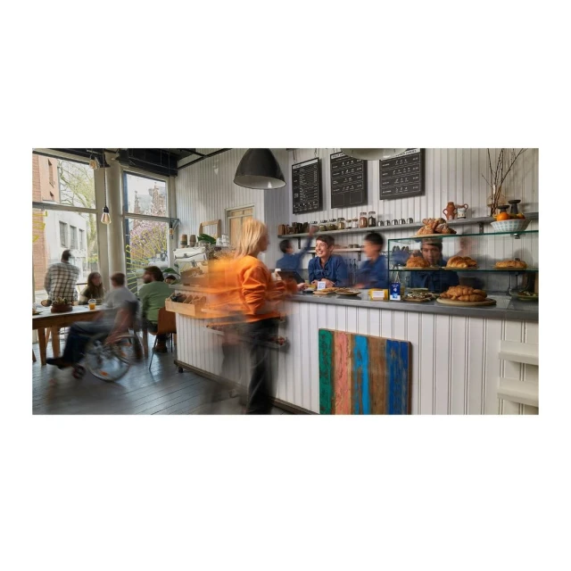 Busy daytime view inside a bakery with patrons at tables and people at the counter, featuring baked goods and menu boards.
