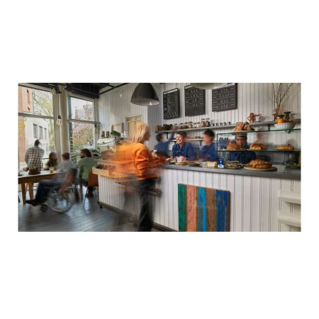 Busy daytime view inside a bakery with patrons at tables and people at the counter, featuring baked goods and menu boards.