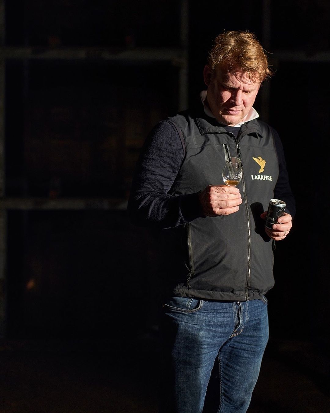 Man in a black vest and blue jeans, holding a glass of whisky and a beverage can, indoors with dim lighting.