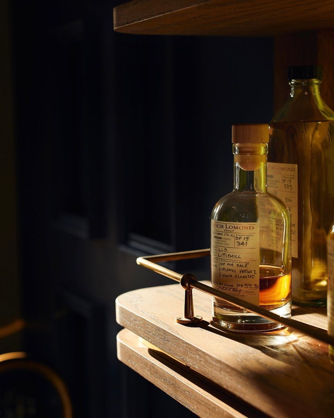 Close-up of two glass bottles on a dark wood surface with brass accents, bathed in warm light against a dark background.