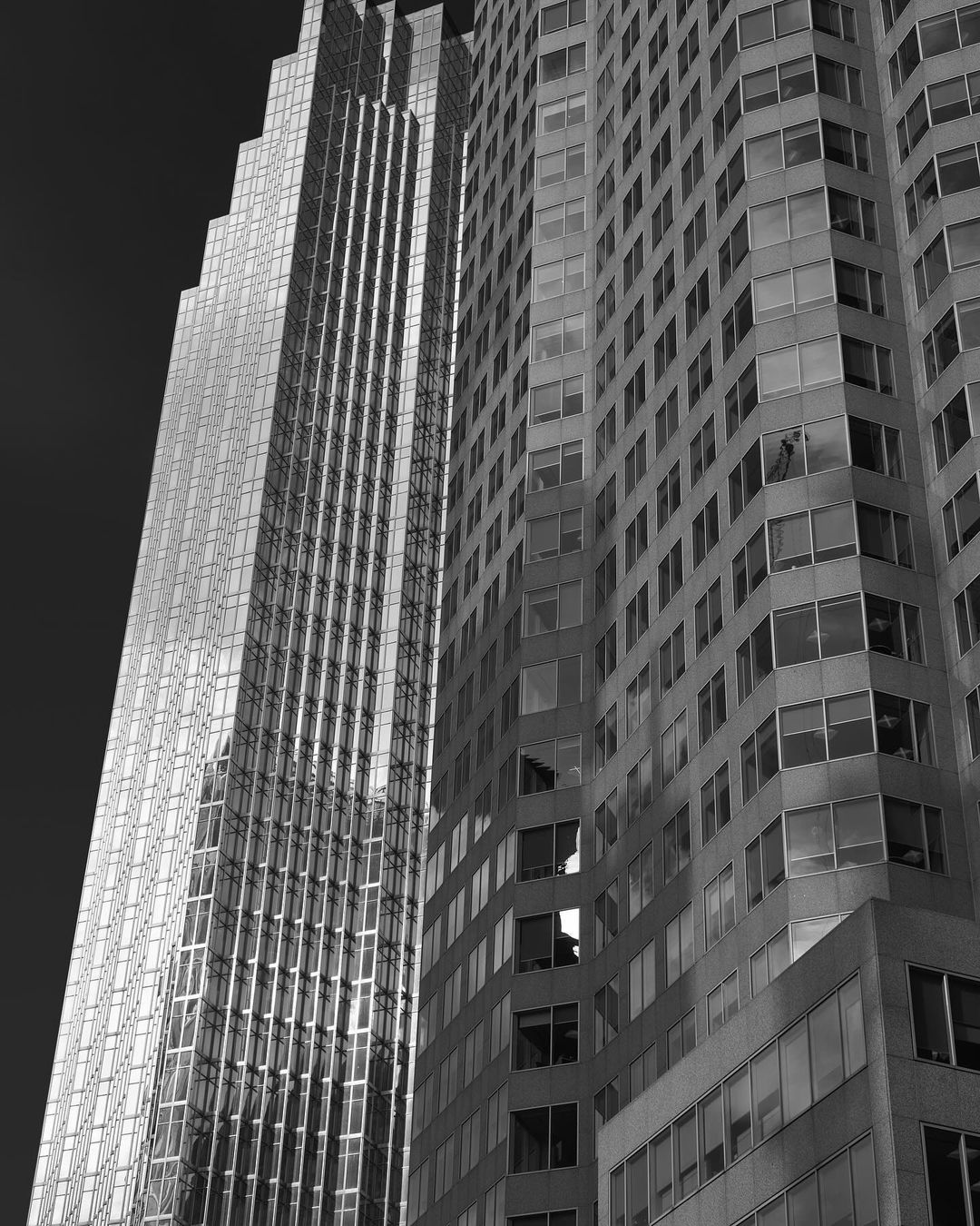 Monochrome photograph of two skyscrapers: one with a smooth, reflective facade and the other with a grid of windows. Urban architecture.