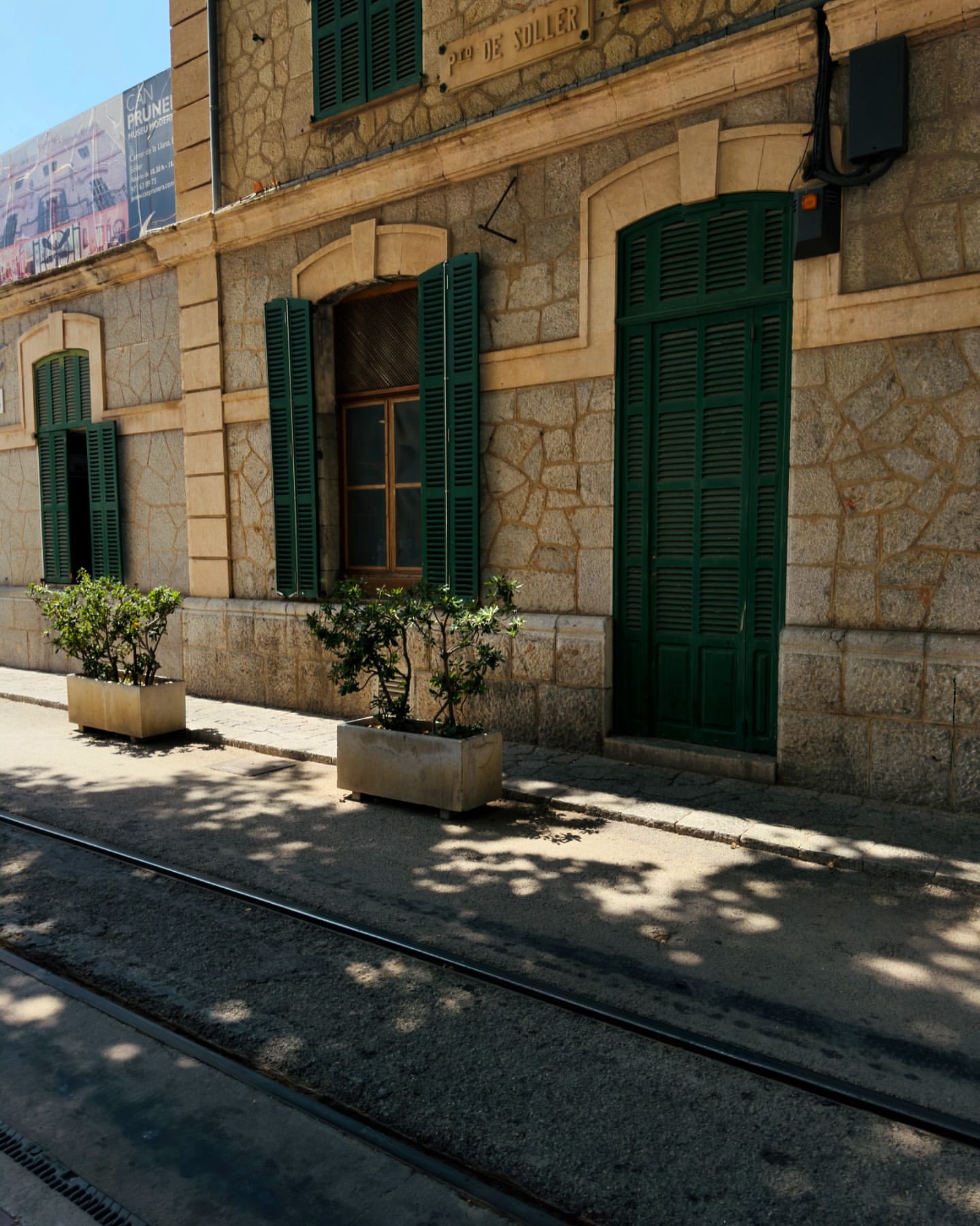 Exterior of a stone building with green shutters and a door, complemented by potted plants on a sunny day, creating a tranquil scene.
