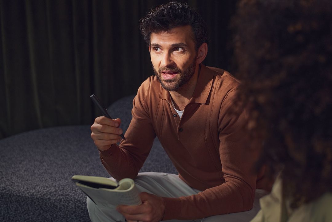 Adult male in a rust-colored shirt talking while holding a pen and notebook, indoor environment, with soft, natural lighting.
