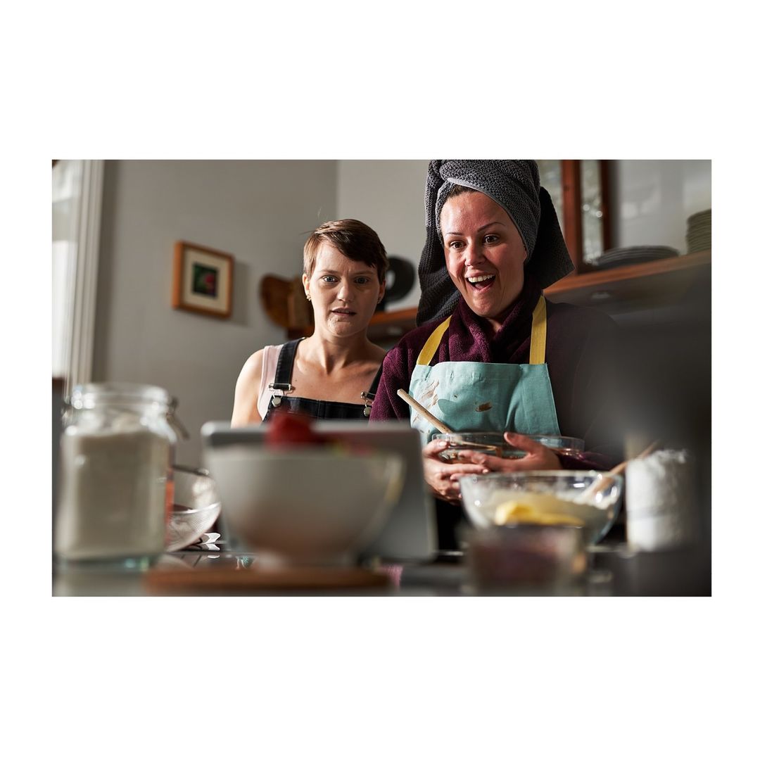 Two adult women in a kitchen prepare food with bowls, ingredients, and tea towels in an indoor, domestic setting with daylight.