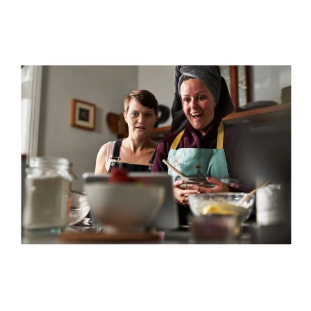 Two adult women in a kitchen prepare food with bowls, ingredients, and tea towels in an indoor, domestic setting with daylight.