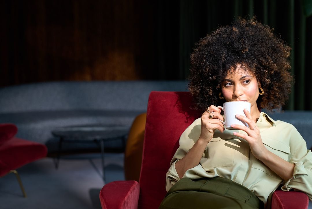 Woman with afro sits in red chair, sipping from white mug in relaxed setting with natural light, wearing blouse and green trousers.