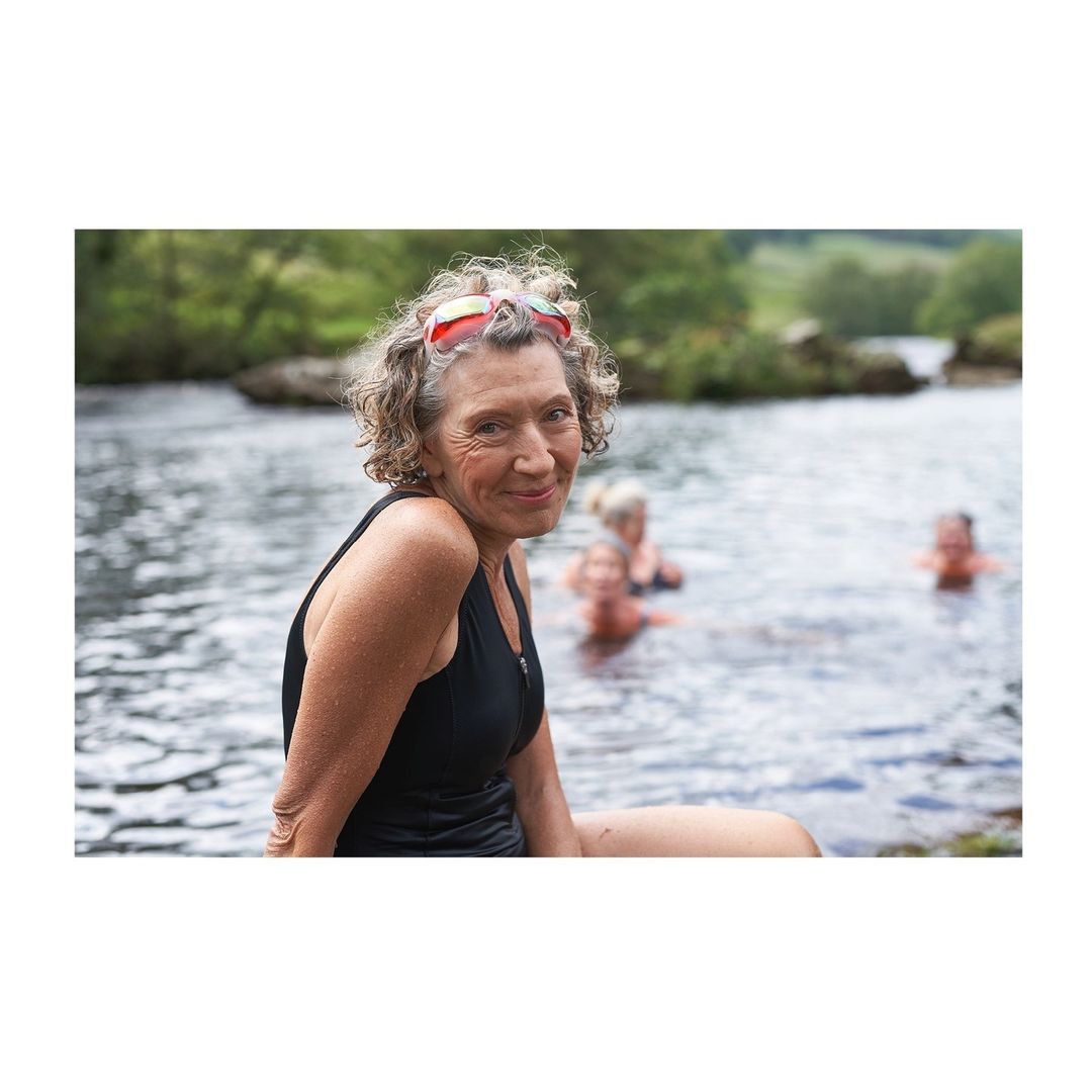 Elderly woman in black swimsuit and swimming goggles poses by a river with other people in the water and trees in the background.