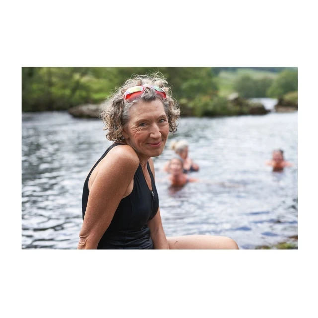 Elderly woman in black swimsuit and swimming goggles poses by a river with other people in the water and trees in the background.