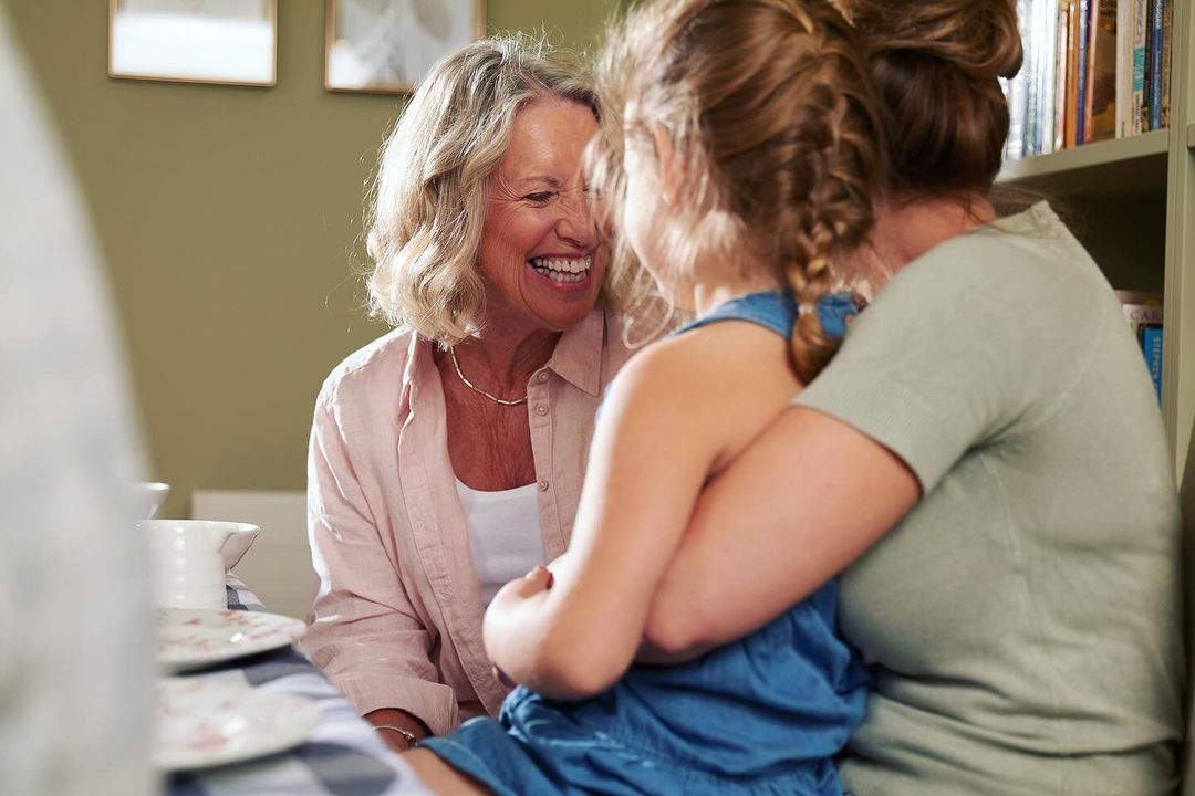 Three generations of women embracing in a cozy indoor setting with a bookshelf and natural light, showcasing family togetherness.