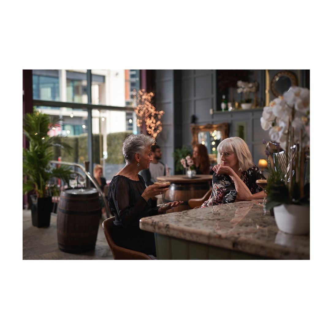 Two adult women sitting at a bar with marble countertop, engaged in conversation in an indoor setting with decorative lighting.
