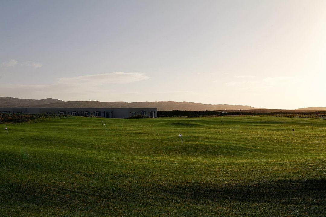 Scenic view of a verdant green golf course with rolling hills, a modern building, and mountains in the soft, warm light of dusk or dawn.