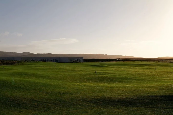 Scenic view of a verdant green golf course with rolling hills, a modern building, and mountains in the soft, warm light of dusk or dawn.
