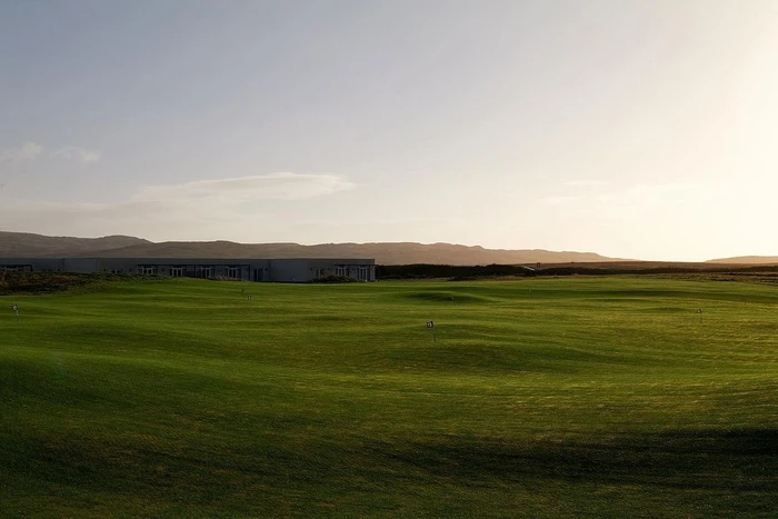 Wide landscape shot of a sprawling green golf course under a pale sky with distant rolling hills and low buildings on the horizon