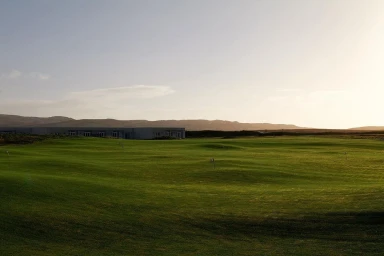 Wide landscape shot of a sprawling green golf course under a pale sky with distant rolling hills and low buildings on the horizon
