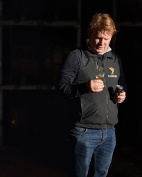 Middle-aged man in a dark gilet and jeans inspecting a tasting glass in a dim interior